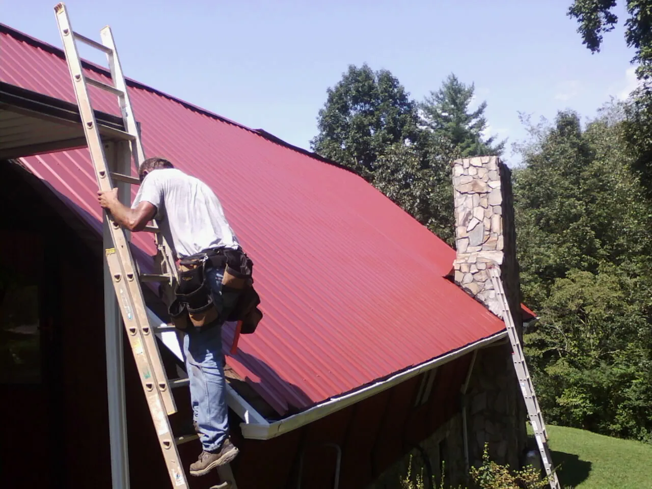 Roofer on a ladder beside a red metal roof and stone chimney — Soffit & Fascia Repair by Roofing Brothers in El Cerrito